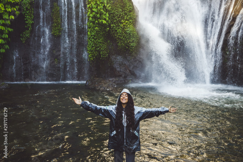 Young happy woman enjoying waterfall in Bali. Wearing grey raincoat from water spray. Travel in Indonesia.
