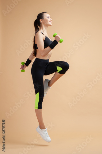 A young brunette woman is engaged in fitness, jumping with dumbbells, raising her knees high, on a peach background.