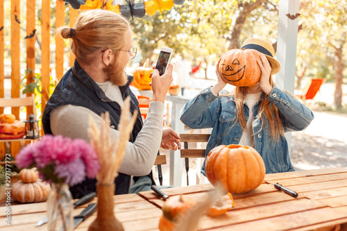 Halloween Preparaton Concept. Young couple sitting at table outdoors making jack-o'-lantern man taking photo of woman with pumpkin