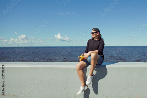 A young man with long hair in denim shorts, sweatshirt is sitting with a yellow skate on the city's quay. photo