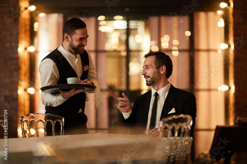 Portrait of mature bearded businessman talking to waiter bringing coffee and smiling happily in luxury restaurant, copy space