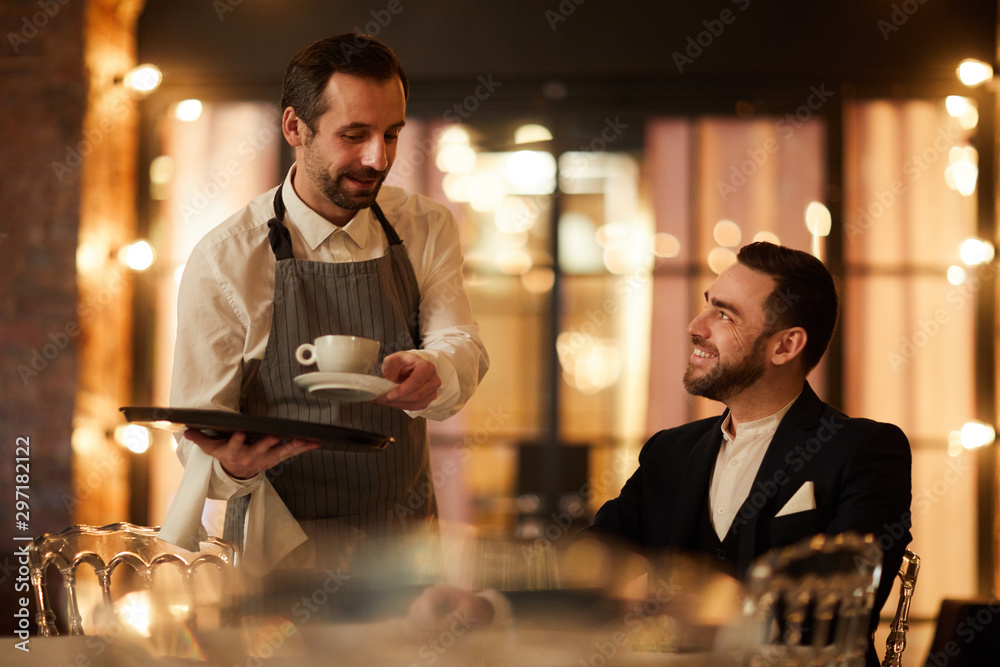Side view portrait of handsome bearded businessman talking to waiter bringing coffee and smiling happily in luxury restaurant