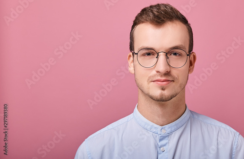 Fototapeta Naklejka Na Ścianę i Meble -  A decent young man in a sky blue shirt and computer glasses stands on a pink background. businessman, employee of the year. Looks straight.