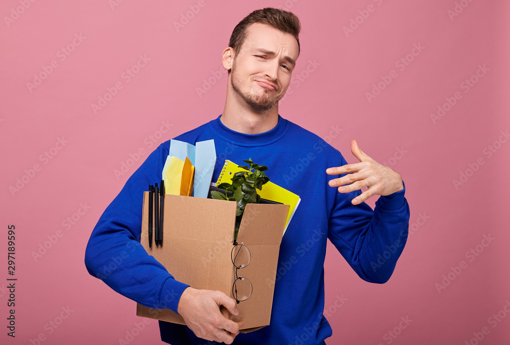 Focus at the box. A cool stylish retro guy in dark sweater is standing hugging a box with green plant, yelow notebook and paper airplane with his both hands. The guy was lose work, but it ok. Focus at the box. A cool stylish retro guy in dark sweater is standing hugging a box with green plant, yelow notebook and paper airplane with his both hands. The guy was lose work, but it ok.