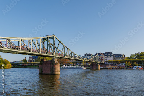 Outdoor sunny view of Eiserner Steg, historical pedestrian Iron Bridge, and promenade on riverside of Main River in sunny day in Frankfurt, Germany. © Peeradontax