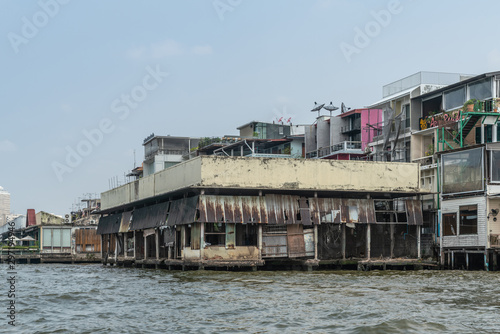 Bangkok city, Thailand - March 17, 2019: Chao Phraya River. Run-down and half demolished warehouse and poor dwellings built on stilts just above water under ligh blue sky. © Klodien