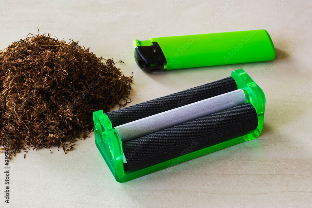 Green cigarette rolling machine, green lighter and pile of tobacco on a white table. Making cigarettes with pipe tobacco at home. Green cigarette rolling machine, green lighter and pile of tobacco on a white table. Making cigarettes with pipe tobacco at home.