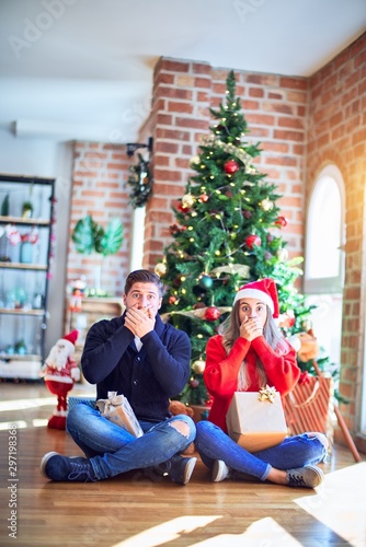 Young couple wearing santa claus hat sitting on the floor around christmas tree at home shocked covering mouth with hands for mistake. Secret concept. © Krakenimages.com