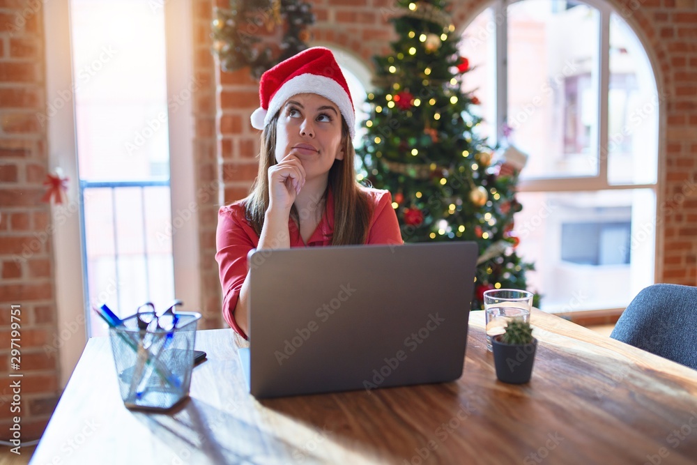 Beautiful woman sitting at the table working with laptop wearing santa claus hat at christmas with hand on chin thinking about question, pensive expression. Smiling with thoughtful face Beautiful woman sitting at the table working with laptop wearing santa claus hat at christmas with hand on chin thinking about question, pensive expression. Smiling with thoughtful face
