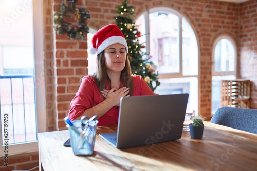 Beautiful woman sitting at the table working with laptop wearing santa claus hat at christmas smiling with hands on chest with closed eyes and grateful gesture on face. Health concept.