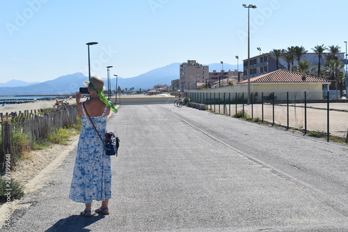 Back of female tourist in long holiday white and blue palm tree print dress taking photo with a mobile phone of the sandy beach from the promenade in south of France. Bright sunny day in Europe. photo