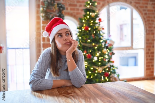 Young beautiful woman wearing santa claus hat at the table at home around christmas decoration thinking looking tired and bored with depression problems with crossed arms.