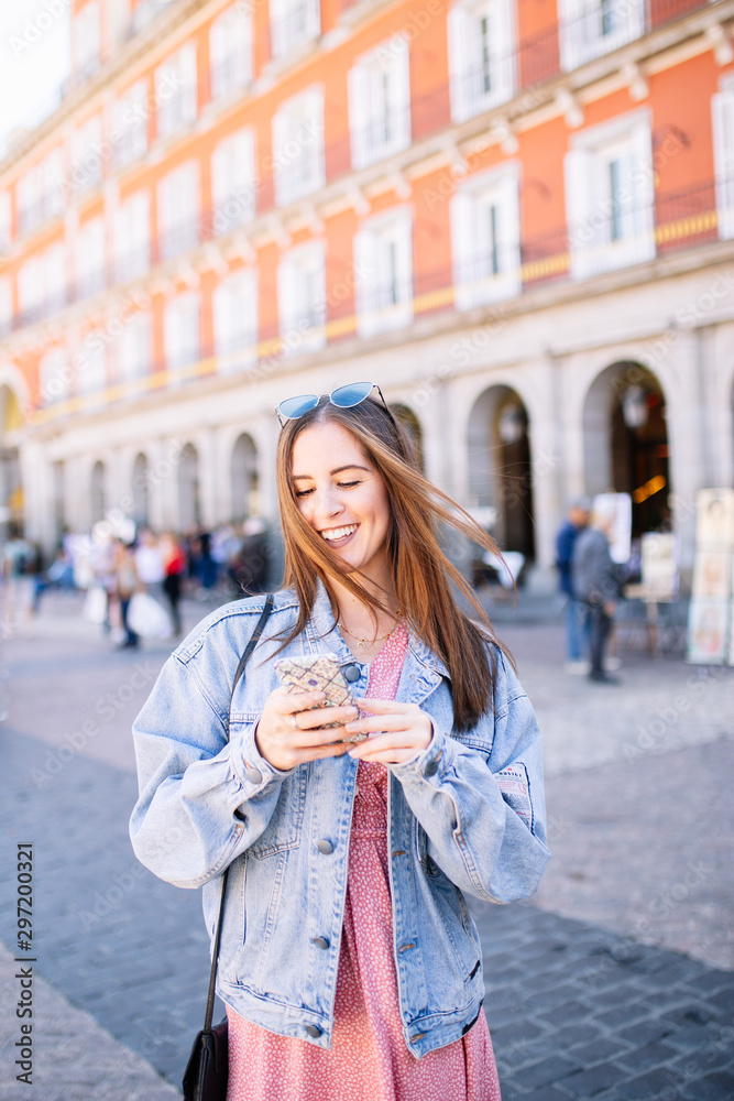 Fototapeta premium Portrait of stylish smiling happy young woman in the street with perfect white teeth looking her mobile. Summer fashion trend, denim jacket, sunglasses, cheerful, positive, laughing. Phone concept.