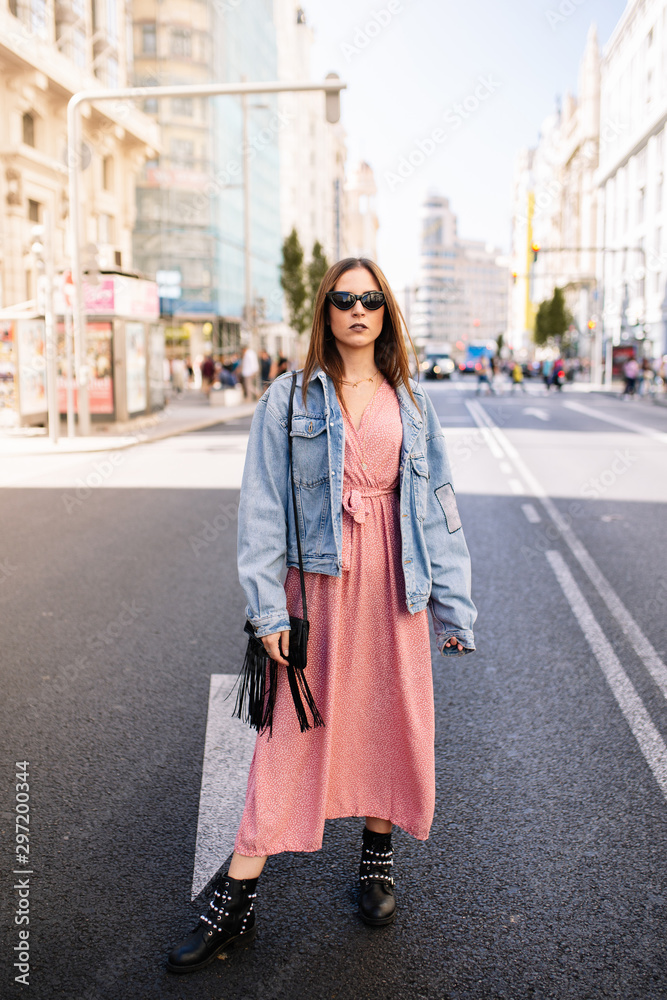 Fototapeta premium Young woman in an pink dress, denim jacket, boots and eye cat sunglasses standing at the famous view of Gran Via main broadway road in the downtown of Madrid, Spain .