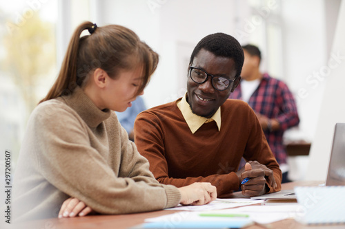 Portrait of smiling African-American student sitting at desk in college and enjoying class with female classmate, copy space