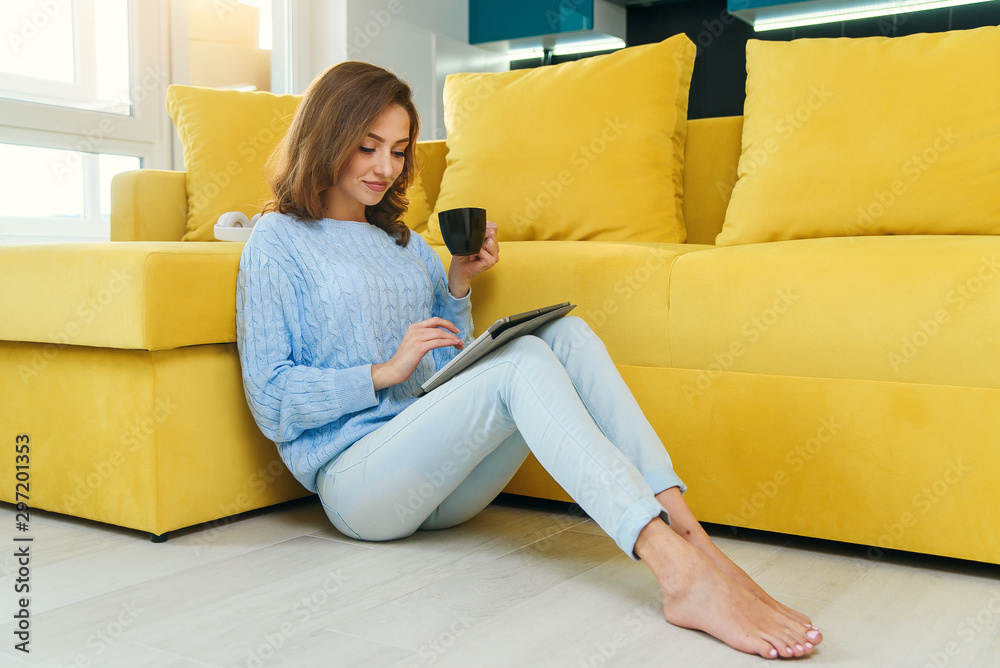 Charming girl holds her tablet computer in hands, sitting on the floor near modern yellow couch and drinks coffee in the modern stylish kitchen. Young cheerful woman having rest at cozy home. Charming girl holds her tablet computer in hands, sitting on the floor near modern yellow couch and drinks coffee in the modern stylish kitchen. Young cheerful woman having rest at cozy home.