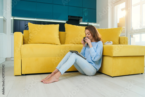 Charming girl holds her tablet computer in hands, sitting on the floor near modern yellow couch and drinks coffee in the modern stylish kitchen. Young cheerful woman having rest at cozy home.