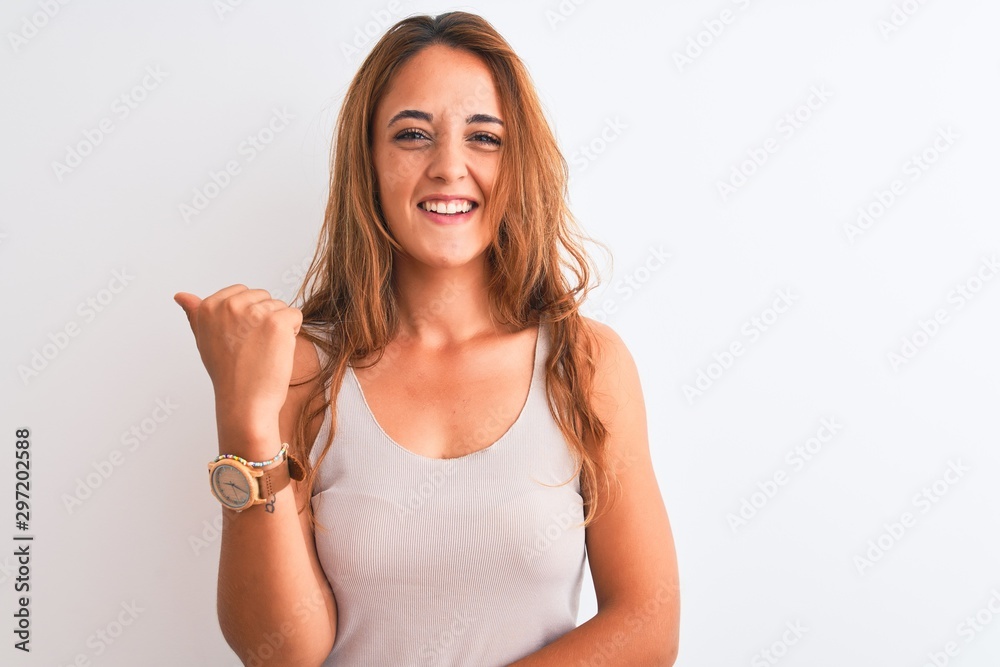 Young redhead woman wearing casual t-shirt stading over white isolated background smiling with happy face looking and pointing to the side with thumb up. Young redhead woman wearing casual t-shirt stading over white isolated background smiling with happy face looking and pointing to the side with thumb up.