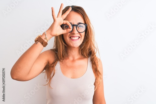Young redhead woman wearing glasses standing over white isolated background doing ok gesture with hand smiling, eye looking through fingers with happy face.