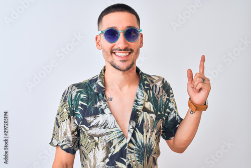 Young handsome man wearing Hawaiian sumer shirt and blue sunglasses over isolated background with a big smile on face, pointing with hand and finger to the side looking at the camera.