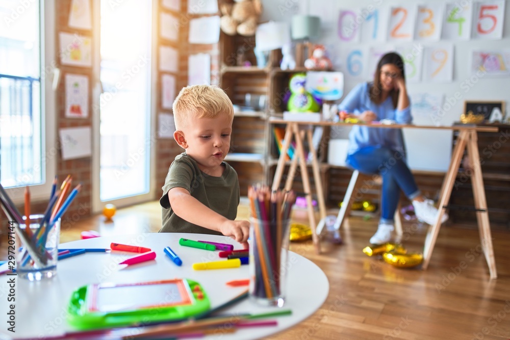 Young caucasian child playing at playschool with teacher. Mother and son at playroom drawing a draw with color pencils, young woman at the background sitting on desk. Young caucasian child playing at playschool with teacher. Mother and son at playroom drawing a draw with color pencils, young woman at the background sitting on desk.