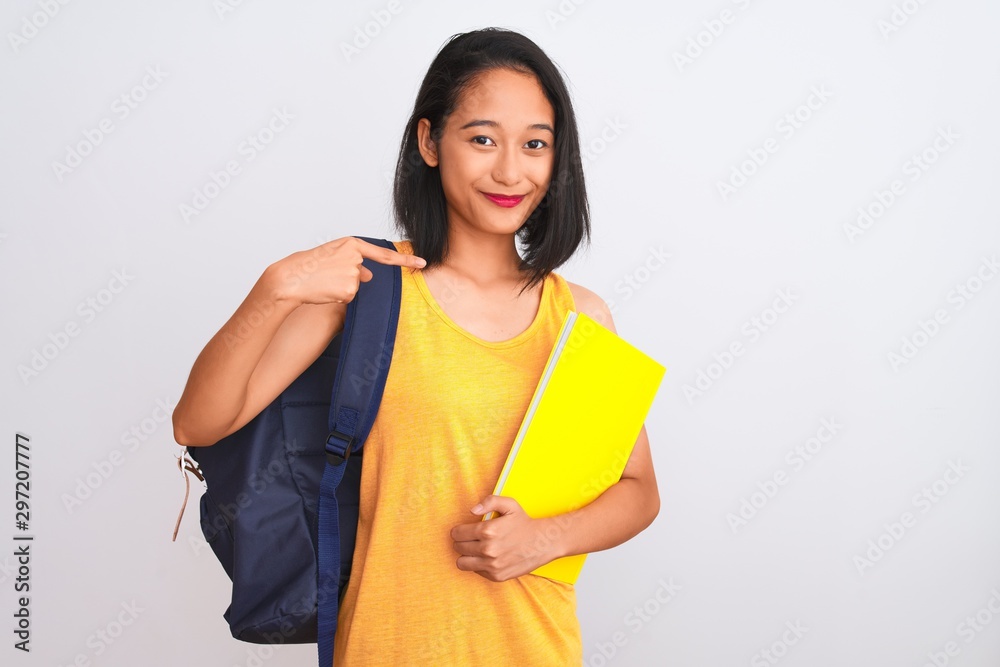 Young chinese student woman wearing backpack holding book over isolated white background with surprise face pointing finger to himself Young chinese student woman wearing backpack holding book over isolated white background with surprise face pointing finger to himself