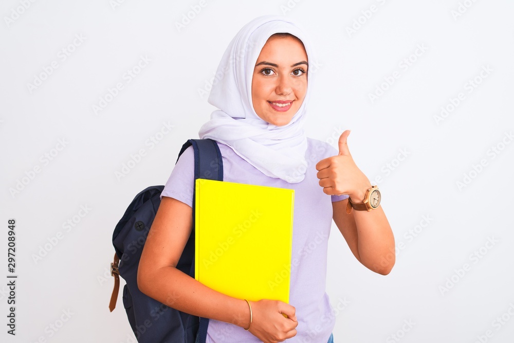 Arab student girl wearing hijab and backpack holding book over isolated white background happy with big smile doing ok sign, thumb up with fingers, excellent sign Arab student girl wearing hijab and backpack holding book over isolated white background happy with big smile doing ok sign, thumb up with fingers, excellent sign