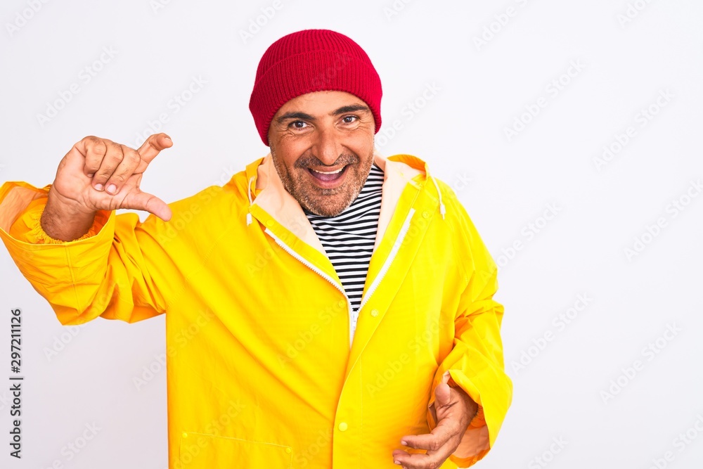 Middle age man wearing rain coat and woolen hat standing over isolated white background smiling and confident gesturing with hand doing small size sign with fingers looking and the camera Middle age man wearing rain coat and woolen hat standing over isolated white background smiling and confident gesturing with hand doing small size sign with fingers looking and the camera