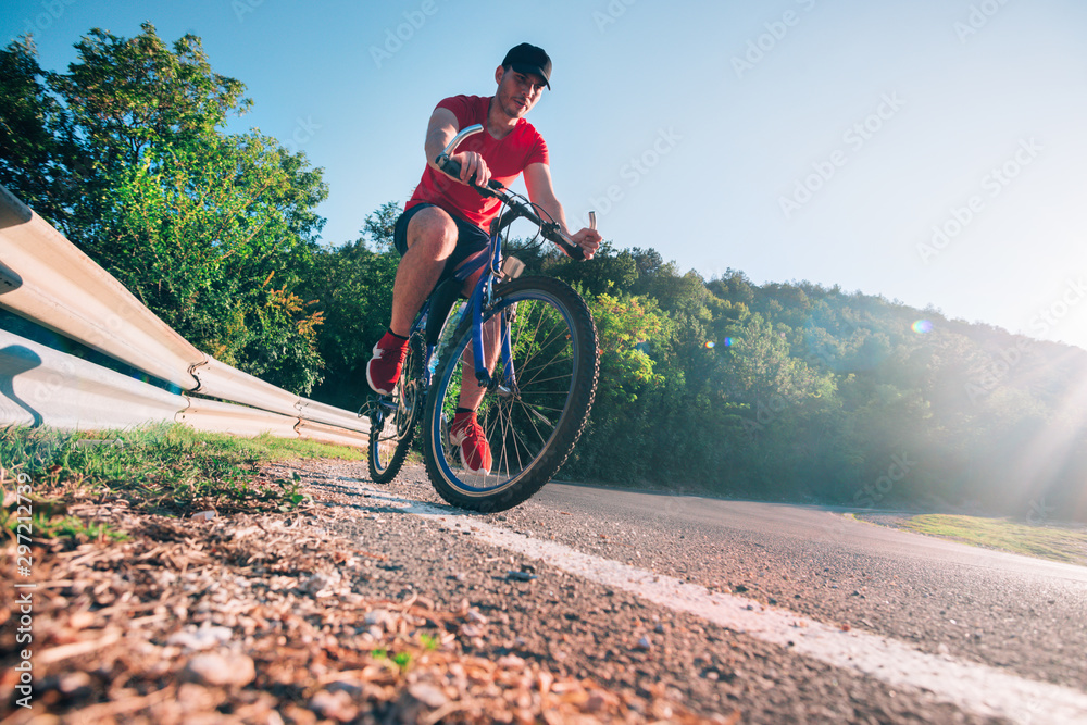 Fototapeta premium Fit male biker cyclist riding his bike cycle on an asphalt road at sunset while the sun sets through his wheel.