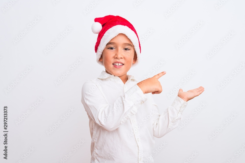 Beautiful kid boy wearing Christmas Santa hat standing over isolated white background amazed and smiling to the camera while presenting with hand and pointing with finger. Beautiful kid boy wearing Christmas Santa hat standing over isolated white background amazed and smiling to the camera while presenting with hand and pointing with finger.
