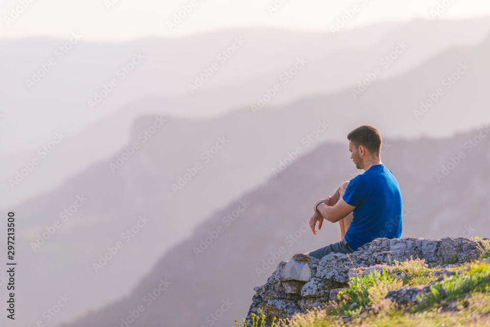 Naklejka premium Adventurous man sitting on top of a mountain and enjoying the beautiful view, while looking downhill at the blue river and amazing mountain line..