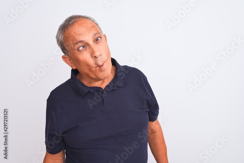 Senior grey-haired man wearing black casual polo standing over isolated white background making fish face with lips, crazy and comical gesture. Funny expression.