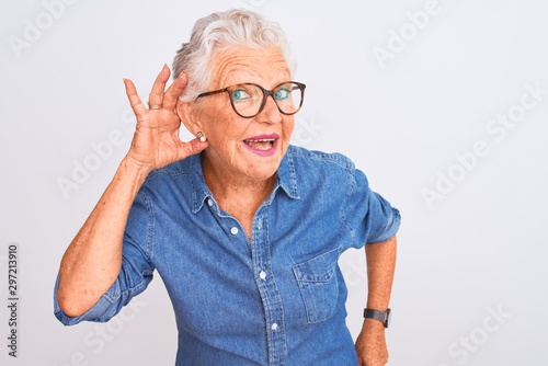 Obraz na płótnie Senior grey-haired woman wearing denim shirt and glasses over isolated white background smiling with hand over ear listening an hearing to rumor or gossip
