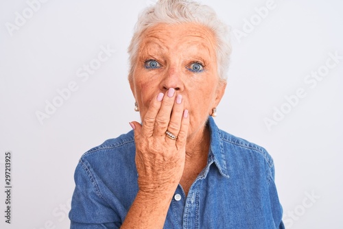 Senior grey-haired woman wearing casual denim shirt standing over isolated white background cover mouth with hand shocked with shame for mistake, expression of fear, scared in silence, secret concept