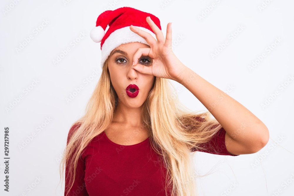 Young beautiful woman wearing Christmas Santa hat over isolated white background doing ok gesture shocked with surprised face, eye looking through fingers. Unbelieving expression. Young beautiful woman wearing Christmas Santa hat over isolated white background doing ok gesture shocked with surprised face, eye looking through fingers. Unbelieving expression.