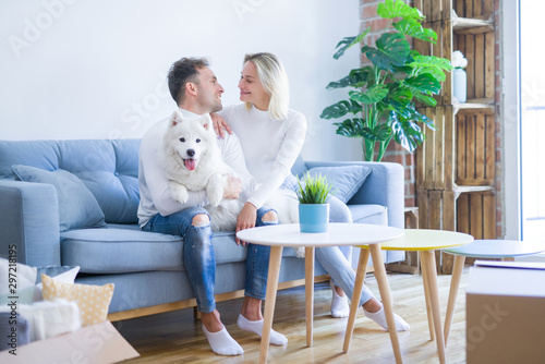 Young beautiful couple with dog sitting on the sofa at new home around cardboard boxes