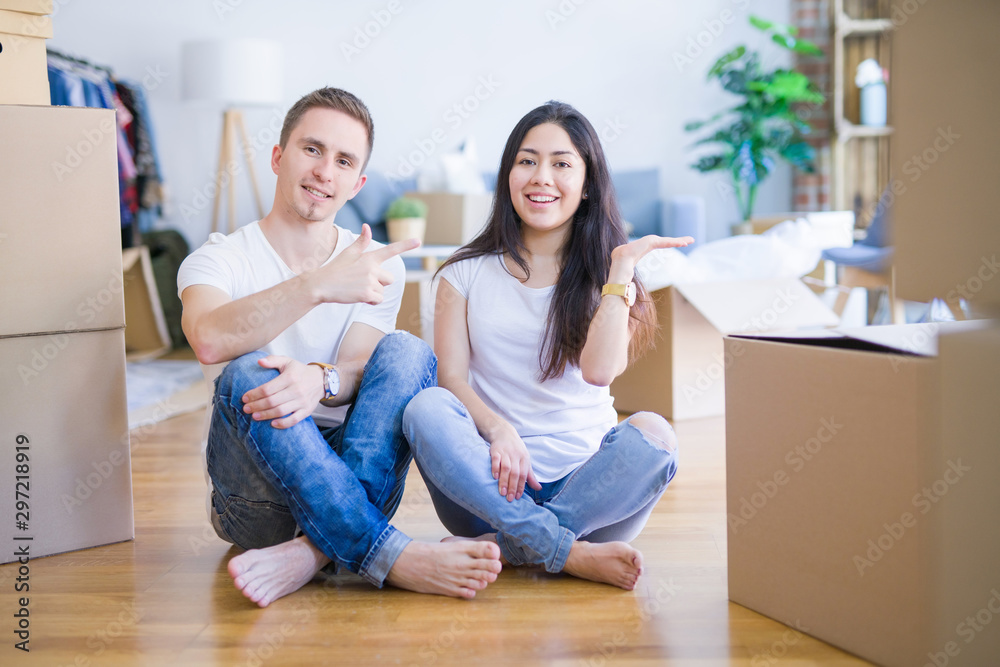 Young beautiful couple sitting on the floor at new home around cardboard boxes amazed and smiling to the camera while presenting with hand and pointing with finger. Young beautiful couple sitting on the floor at new home around cardboard boxes amazed and smiling to the camera while presenting with hand and pointing with finger.