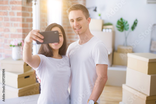 Young beautiful couple standing using smartphone to take a selfie at new home around cardboard boxes