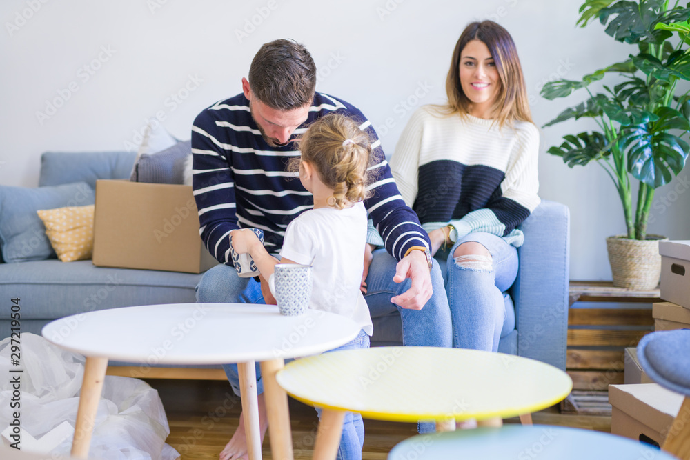 Beautiful family, parents sitting on the sofa drinking coffee looking his kid playing at new home around cardboard boxes Beautiful family, parents sitting on the sofa drinking coffee looking his kid playing at new home around cardboard boxes