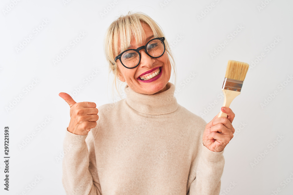 Middle age painter woman holding paint brush standing over isolated white background happy with big smile doing ok sign, thumb up with fingers, excellent sign Middle age painter woman holding paint brush standing over isolated white background happy with big smile doing ok sign, thumb up with fingers, excellent sign