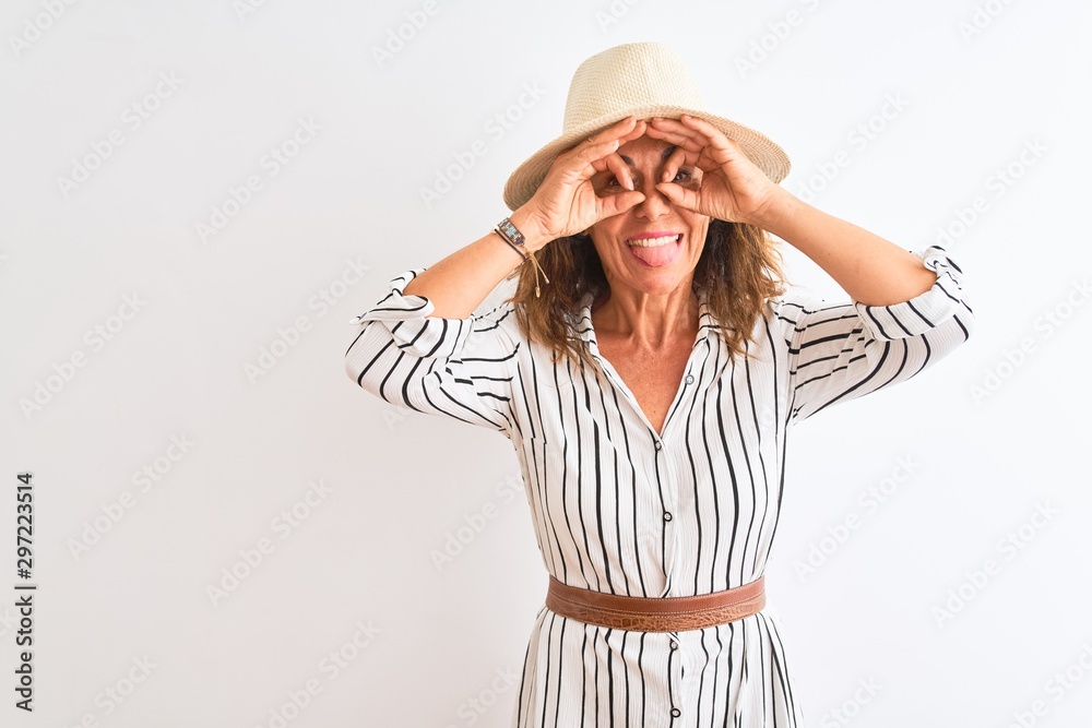 Middle age businesswoman wearing striped dress and hat over isolated white background doing ok gesture like binoculars sticking tongue out, eyes looking through fingers. Crazy expression. Middle age businesswoman wearing striped dress and hat over isolated white background doing ok gesture like binoculars sticking tongue out, eyes looking through fingers. Crazy expression.