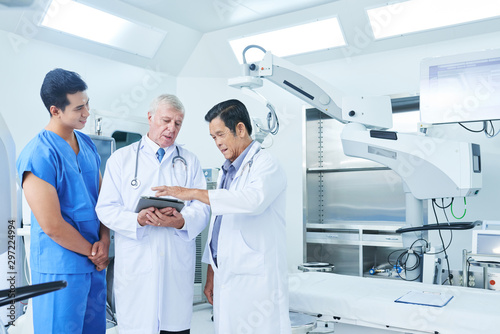 Multi-ethnic general practitioners and surgeon discussing patients data on the screen of tablet computer in operating theatre