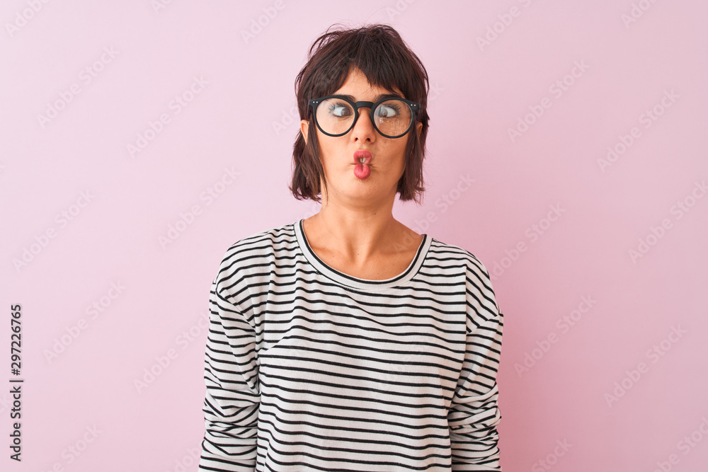 Young beautiful woman wearing striped t-shirt and glasses over isolated pink background making fish face with lips, crazy and comical gesture. Funny expression.