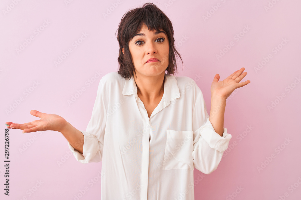 Young beautiful woman wearing white shirt standing over isolated pink background clueless and confused expression with arms and hands raised. Doubt concept. Young beautiful woman wearing white shirt standing over isolated pink background clueless and confused expression with arms and hands raised. Doubt concept.