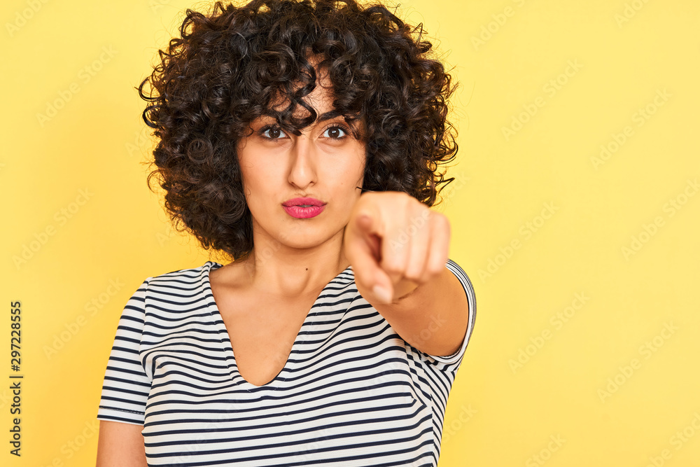 Young arab woman with curly hair wearing striped dress over isolated yellow background pointing with finger to the camera and to you, hand sign, positive and confident gesture from the front Young arab woman with curly hair wearing striped dress over isolated yellow background pointing with finger to the camera and to you, hand sign, positive and confident gesture from the front