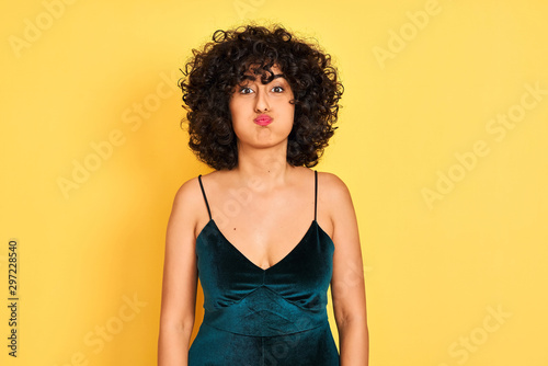 Young arab woman with curly hair wearing elegant dress over isolated yellow background puffing cheeks with funny face. Mouth inflated with air, crazy expression.
