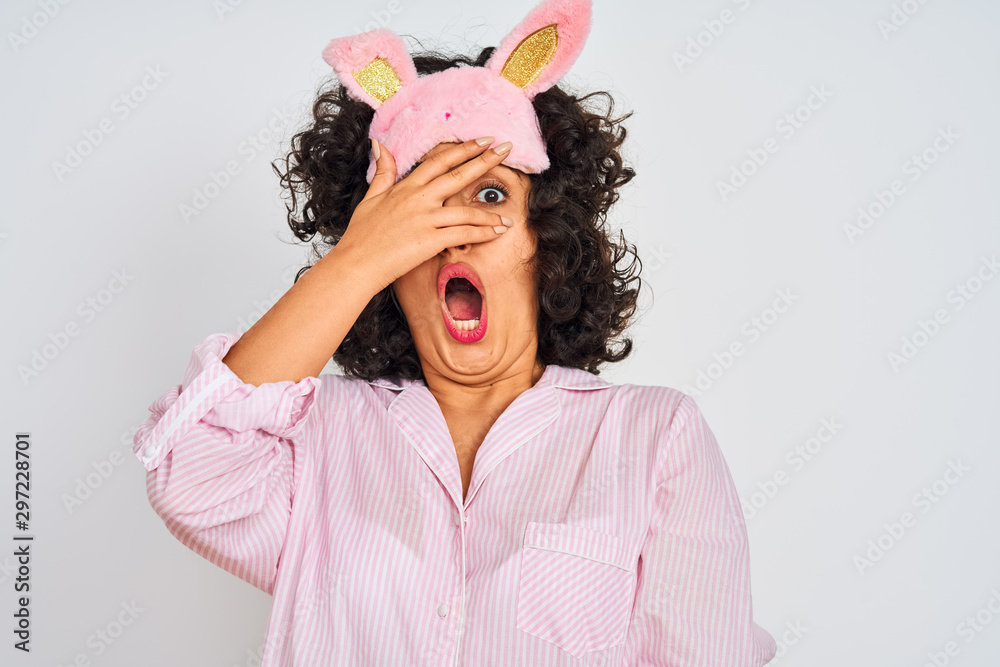 Arab woman with curly hair wearing pajama and sleep mask over isolated white background peeking in shock covering face and eyes with hand, looking through fingers with embarrassed expression.