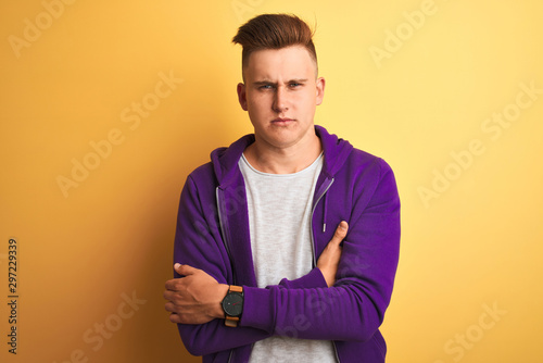 Young handsome man wearing purple sweatshirt standing over isolated yellow background skeptic and nervous, disapproving expression on face with crossed arms. Negative person.