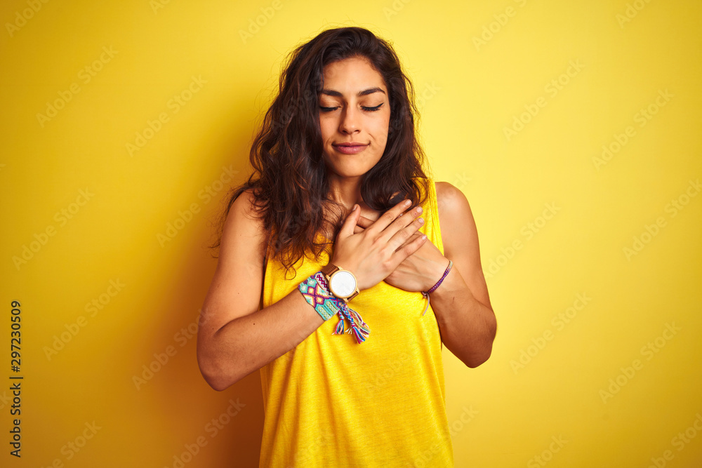Young beautiful woman wearing t-shirt standing over isolated yellow background smiling with hands on chest with closed eyes and grateful gesture on face. Health concept. Young beautiful woman wearing t-shirt standing over isolated yellow background smiling with hands on chest with closed eyes and grateful gesture on face. Health concept.