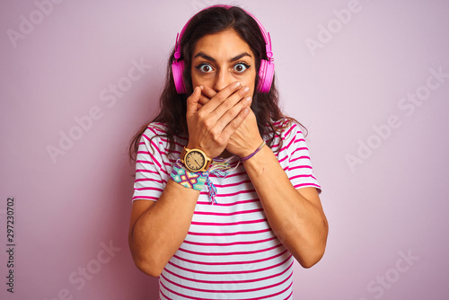 Young beautiful woman listening to music using headphones over isolated pink background shocked covering mouth with hands for mistake. Secret concept.